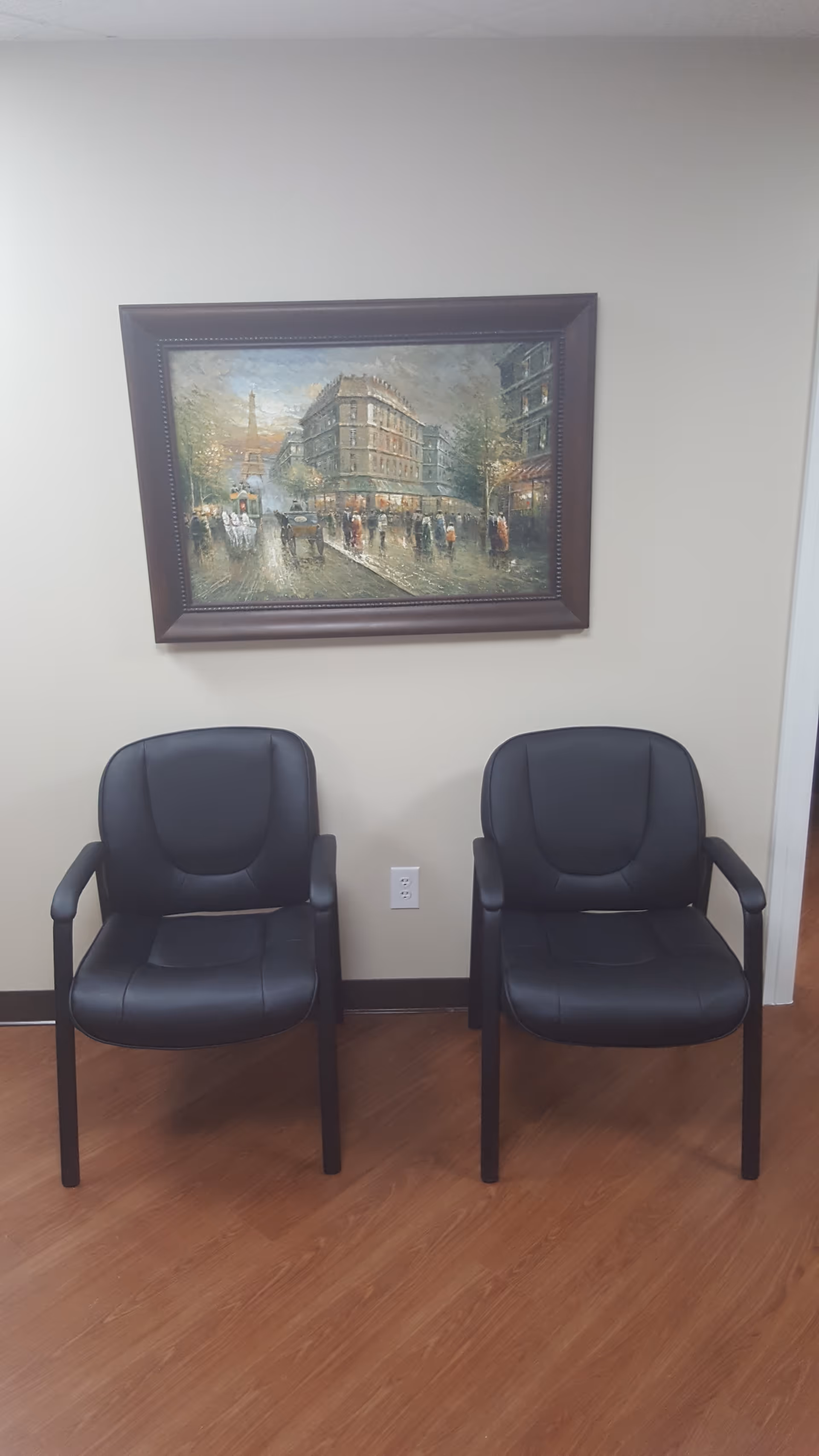 Two black cushioned chairs in a hallway with a framed painting on a beige wall.
