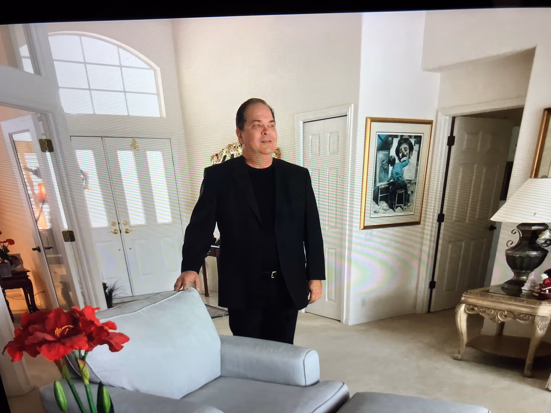 A man in a black suit stands in a bright living room with a sofa.