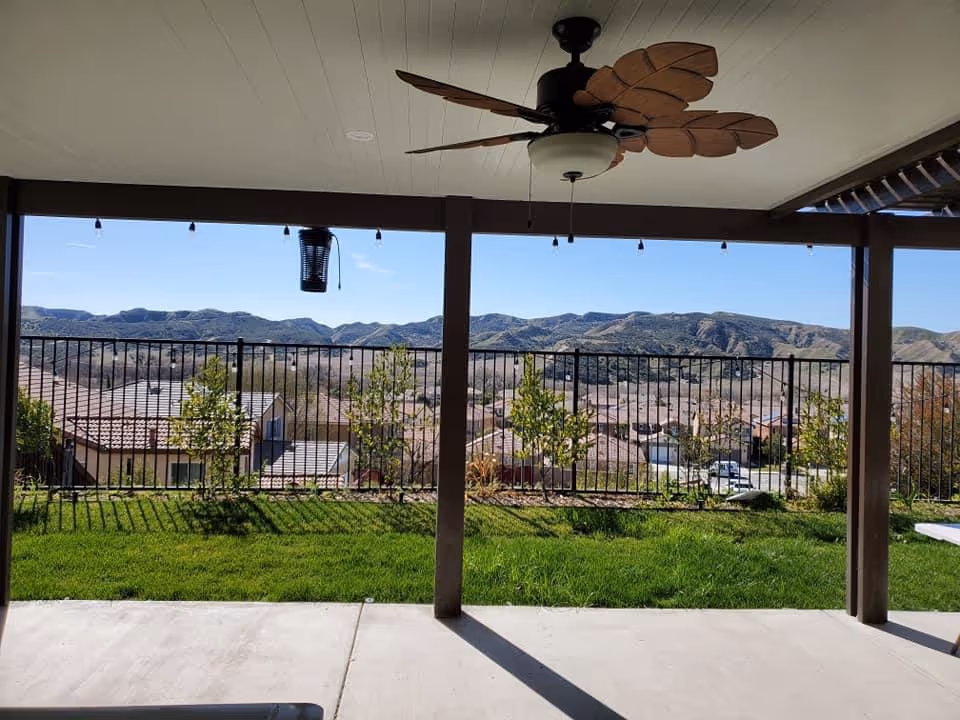 Covered patio overlooking fenced yard, green grass, and distant mountains with a ceiling fan.