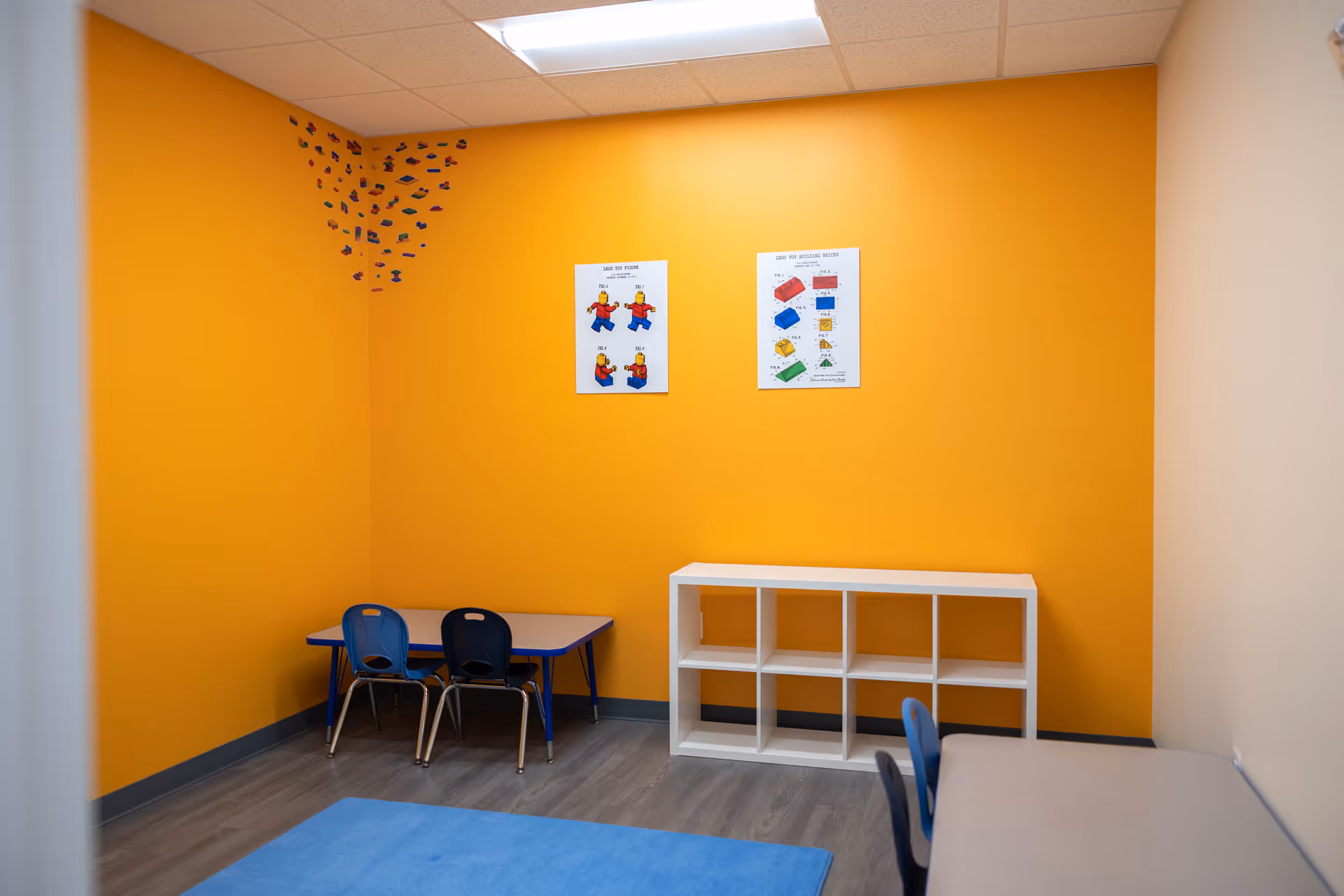 Bright orange activity room with a small table, blue chairs, white cubby shelves, and posters.
