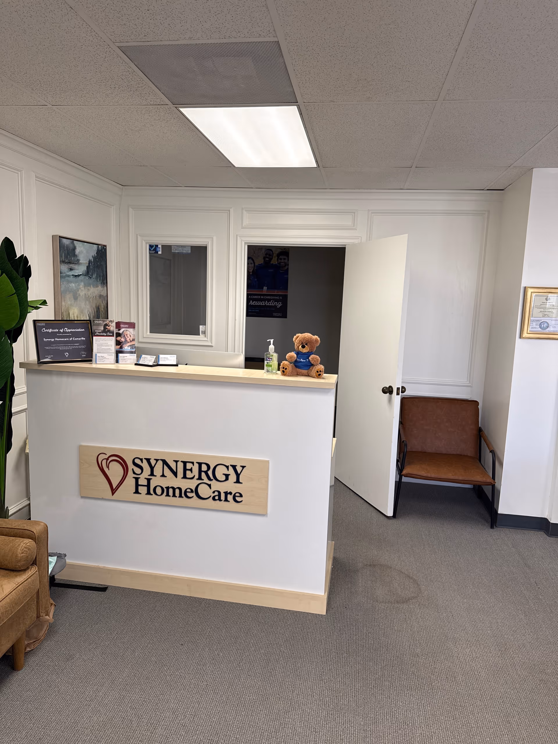 Reception desk with SYNERGY HomeCare sign and teddy bear in a bright lobby.