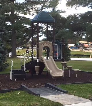 Playground with slide and climbing structure in a grassy, tree-lined outdoor area.