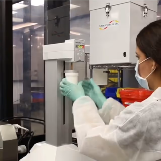 Lab technician in protective gear handles a container beside tall laboratory equipment.