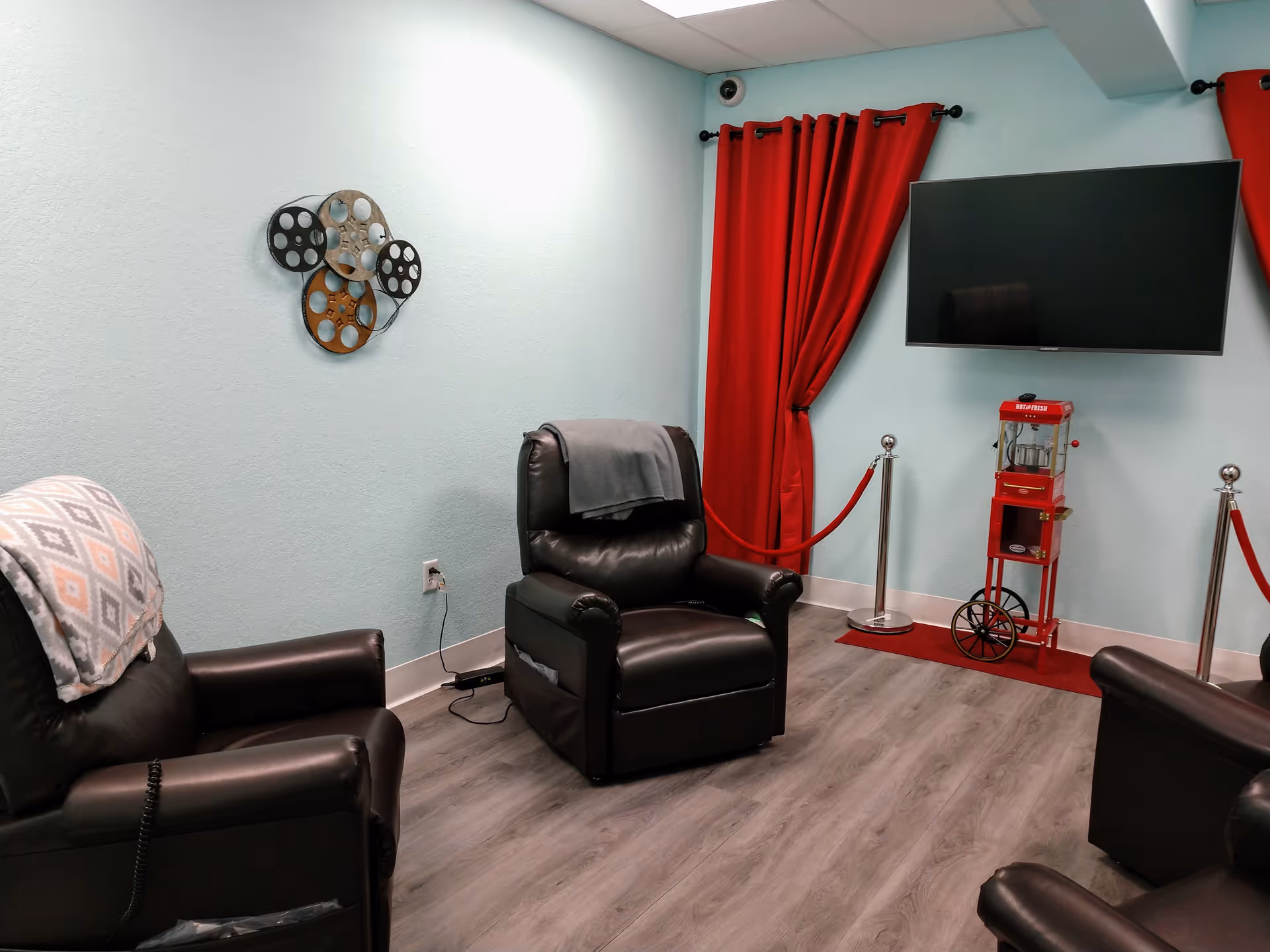 A small senior lounge with teal walls, leather recliners, a wall TV, and red curtains.