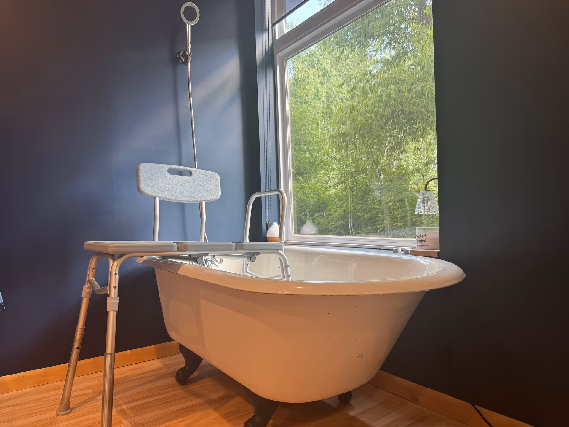 A bathtub with a transfer bench and shower nozzle by a window in a dark bathroom.