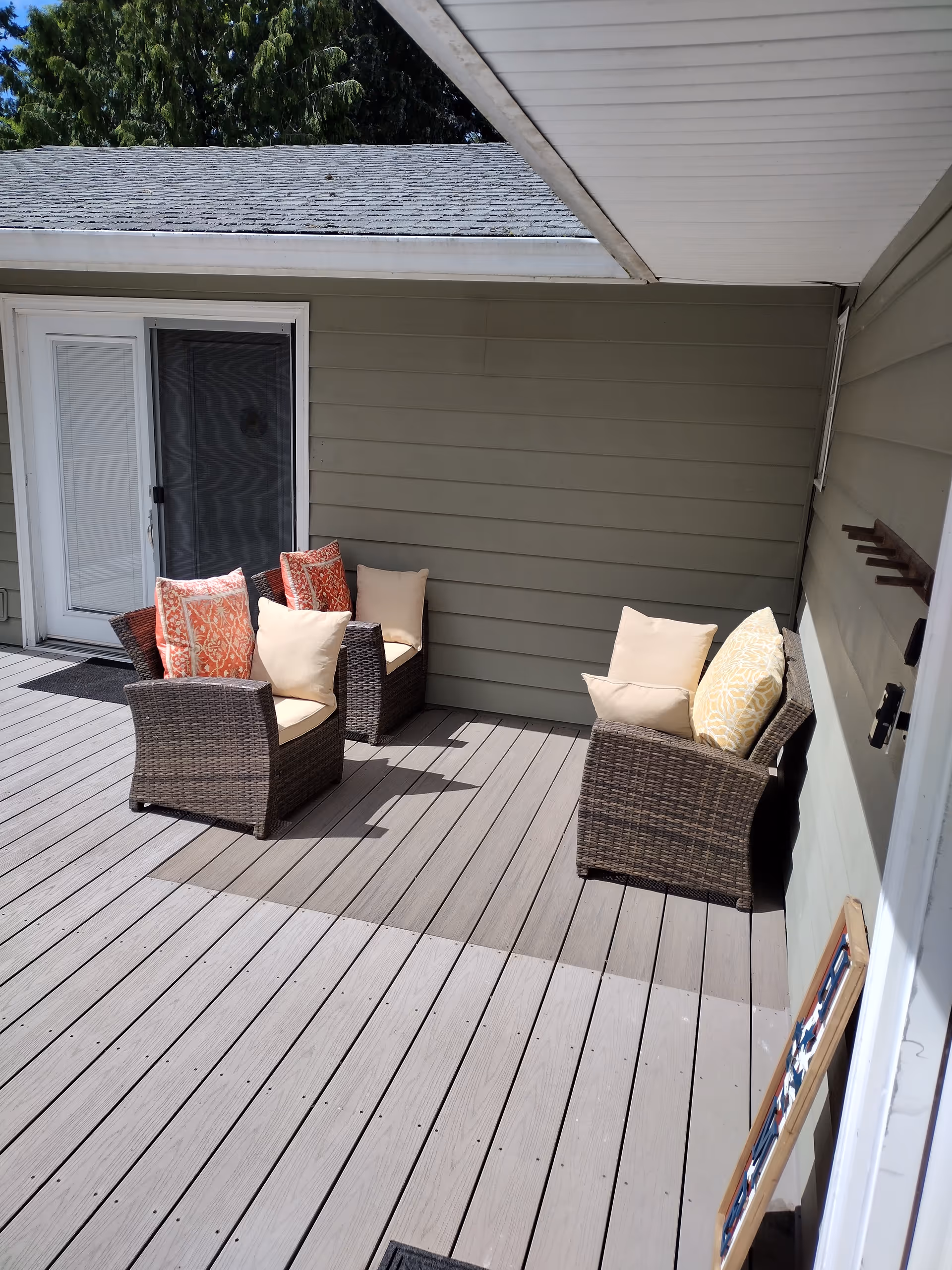 Three wicker chairs with cushions sit on a wooden deck outside a beige house.
