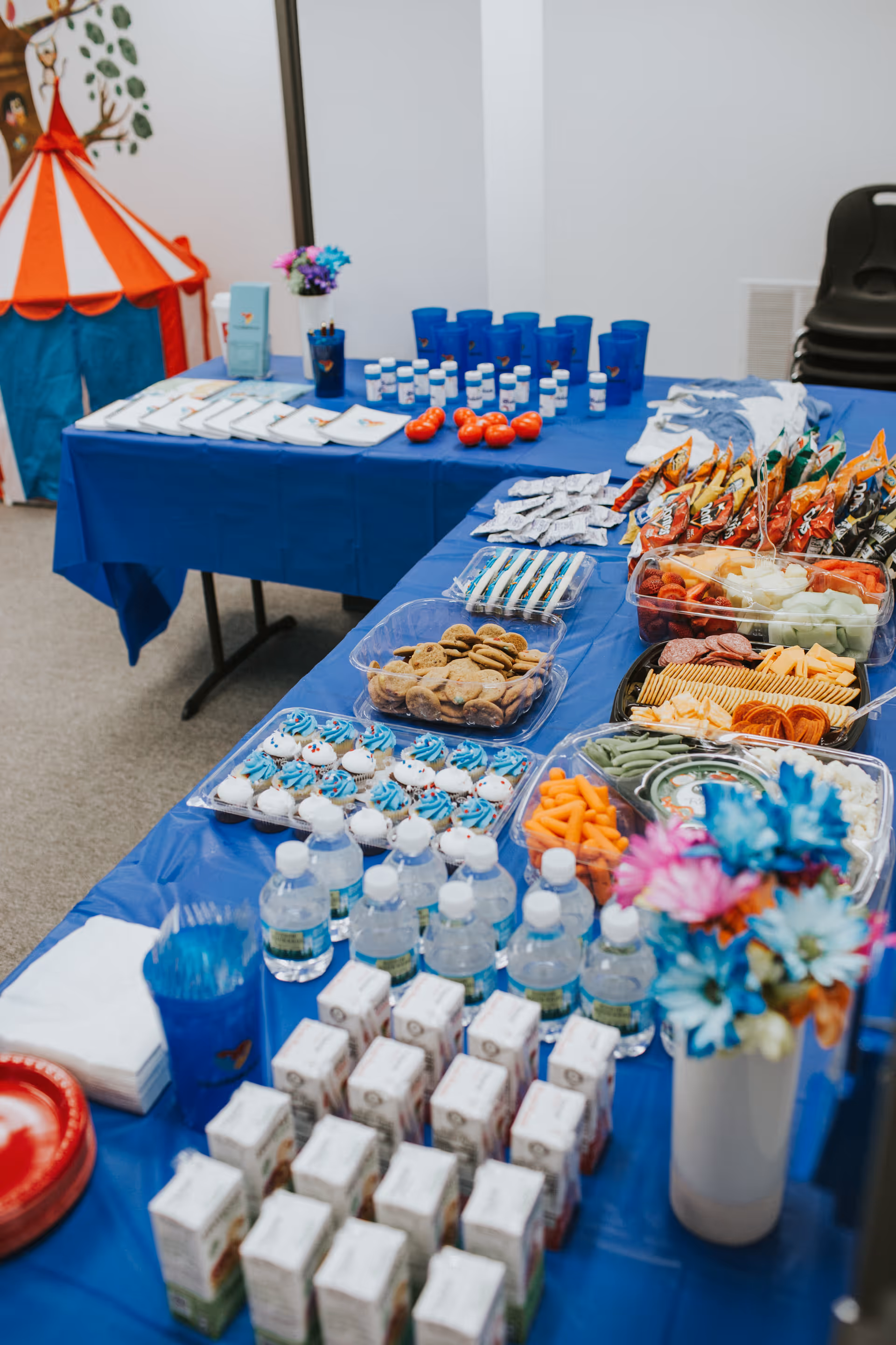 Long blue-draped buffet tables with snacks, drinks, and decorations indoors.