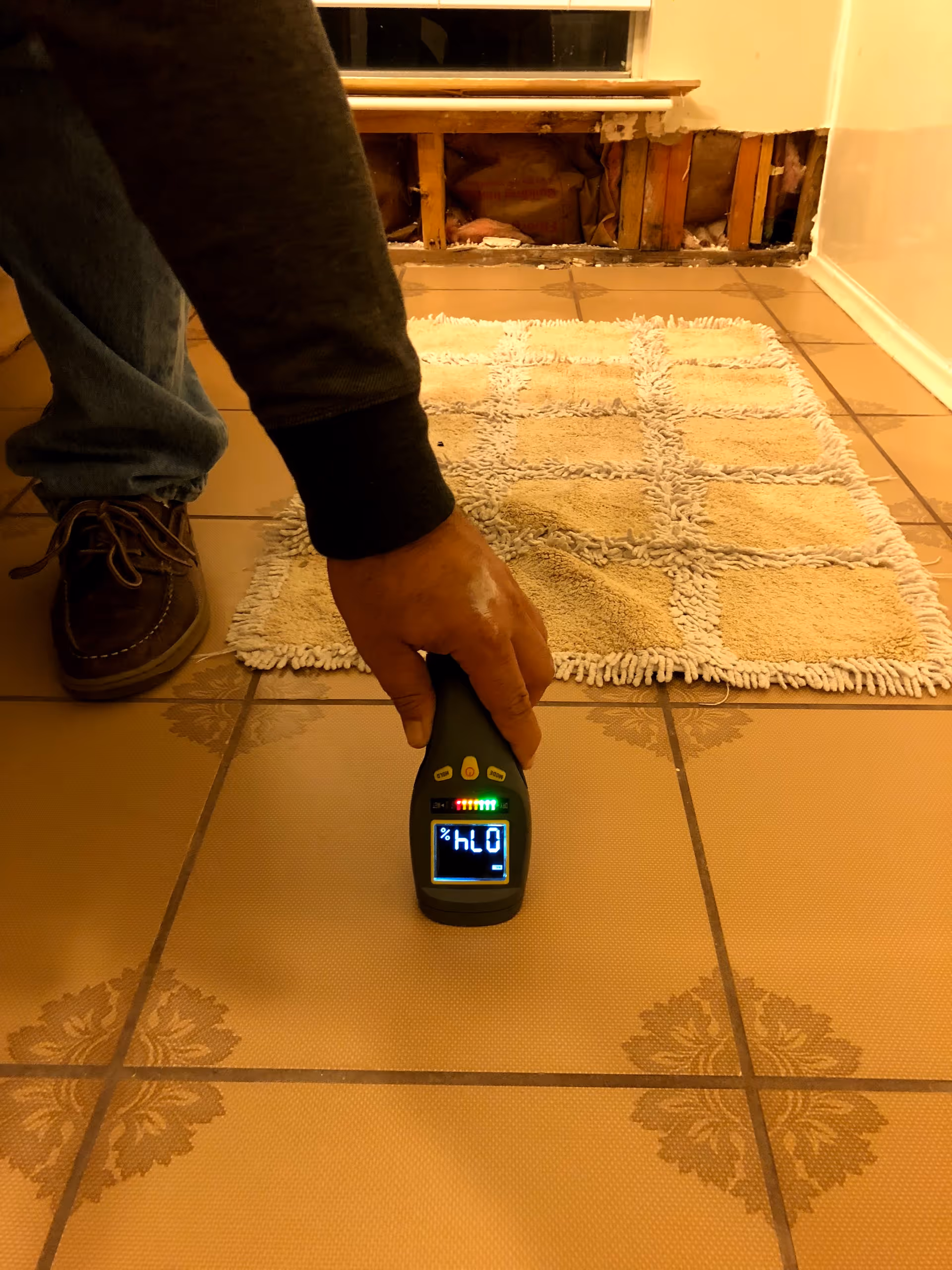 Interior hallway with exposed wall framing and a hand-held moisture meter on tiled floor.