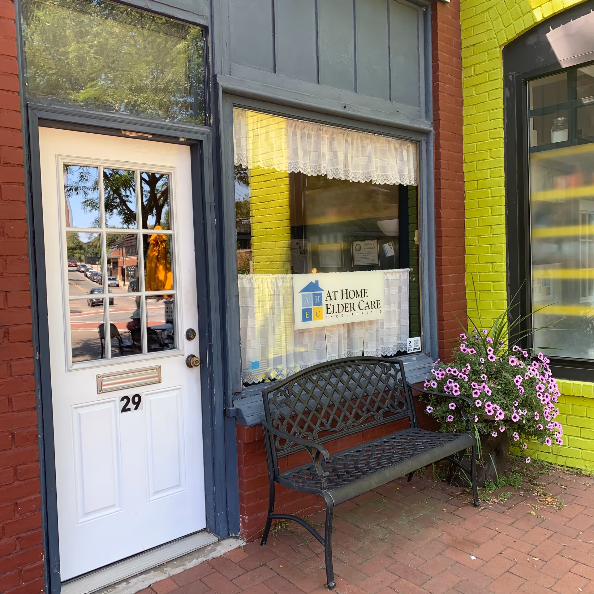 Exterior storefront with white door, lace-window curtain, bench, and a sign reading At Home Elder Care.