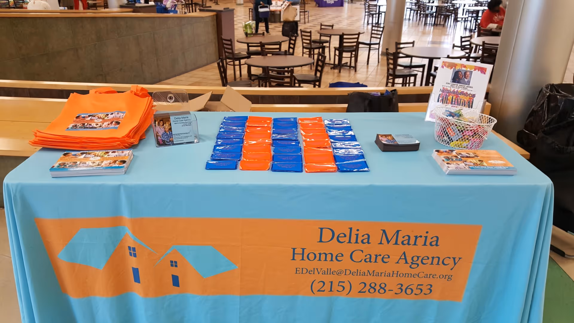 Promotional table for Delia Maria Home Care Agency with brochures in a busy indoor lobby