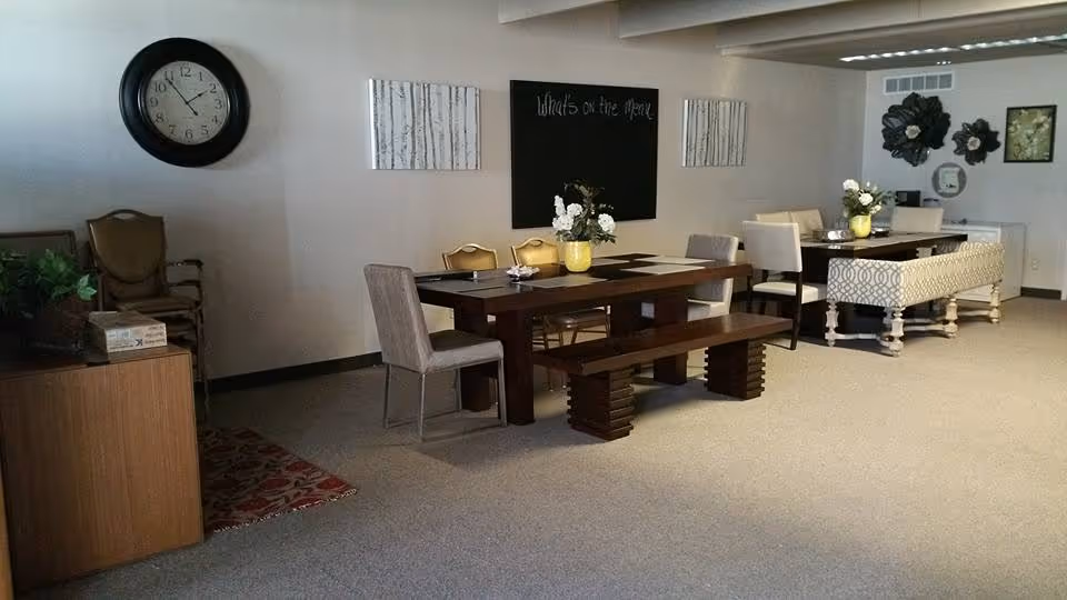 Well-lit senior living common area featuring a long dining table with chairs and a wall clock.
