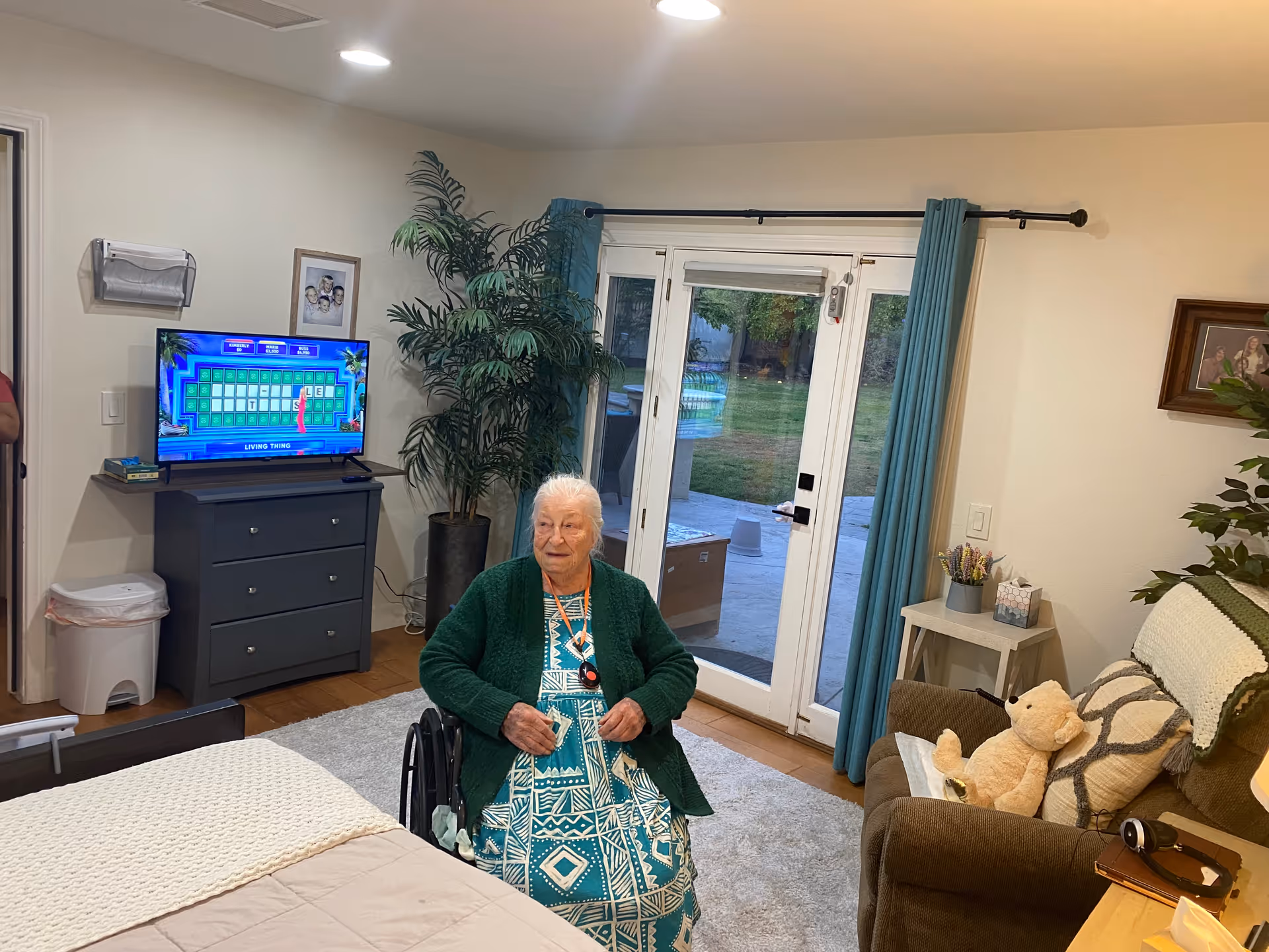 Elderly woman in wheelchair sits in a cozy living room with TV and patio doors.
