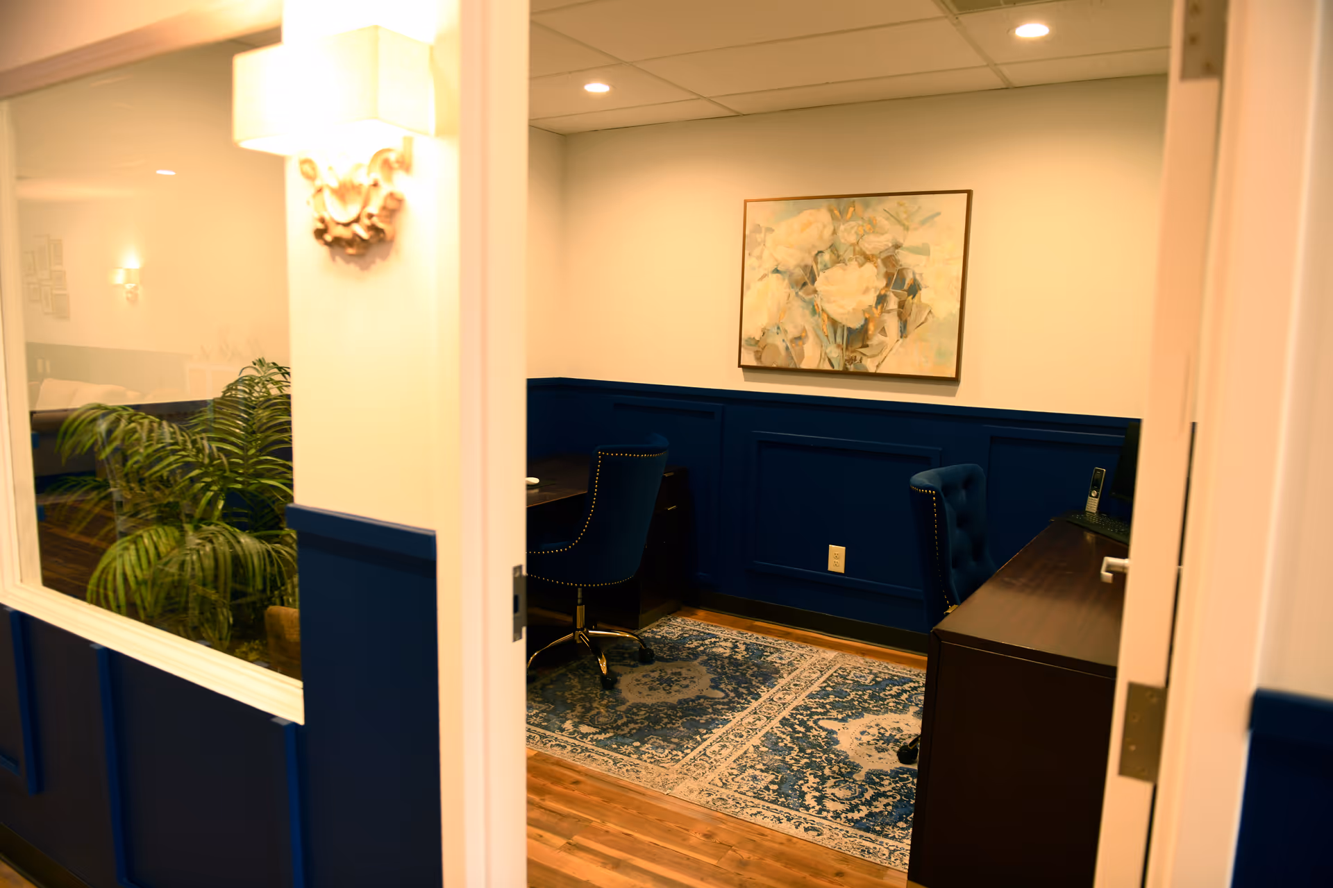 An interior office nook with blue wainscoting, desk, chairs, plant, rug, and artwork.