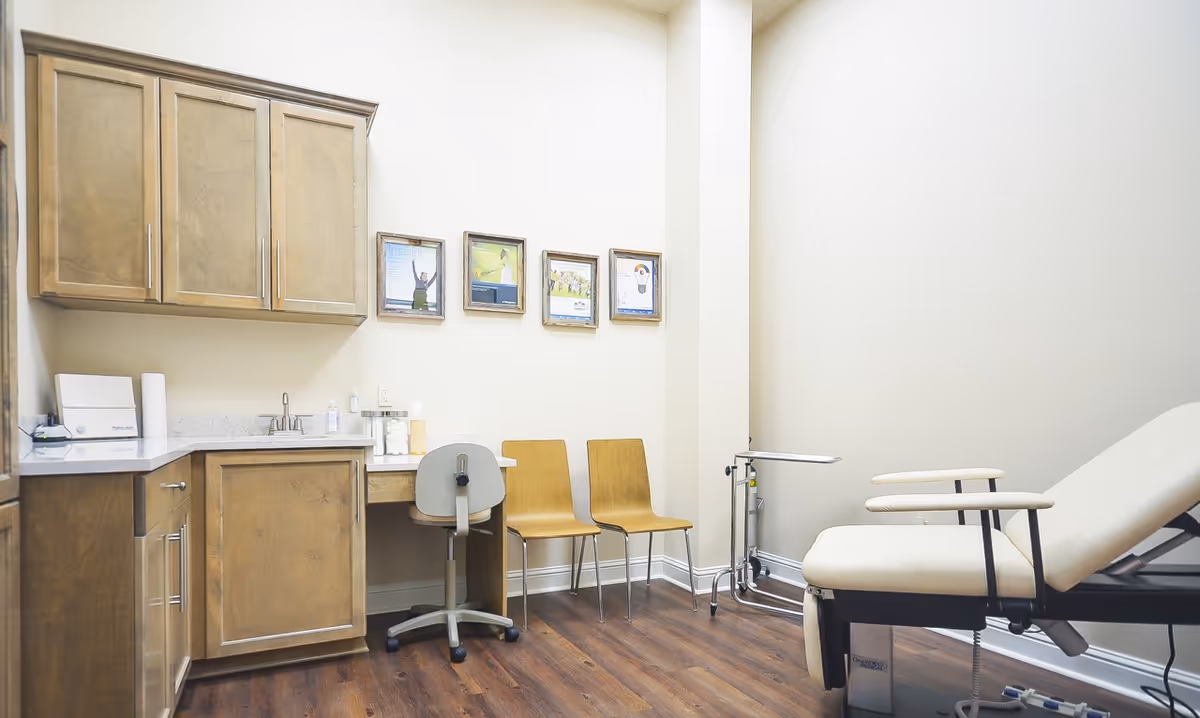 Clinic-style examination room with wooden cabinets, counter, chairs, and examination chair.