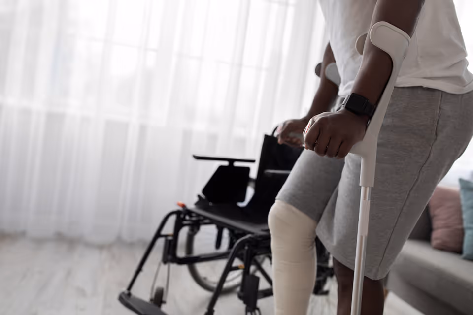 A person uses a white forearm crutch beside a wheelchair in a bright living room.