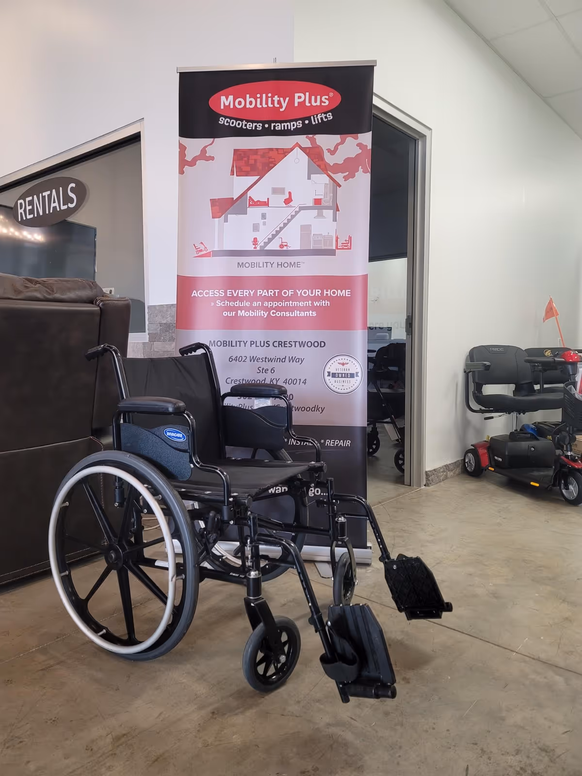 Wheelchair displayed indoors beside Mobility Plus banner in a showroom setting.