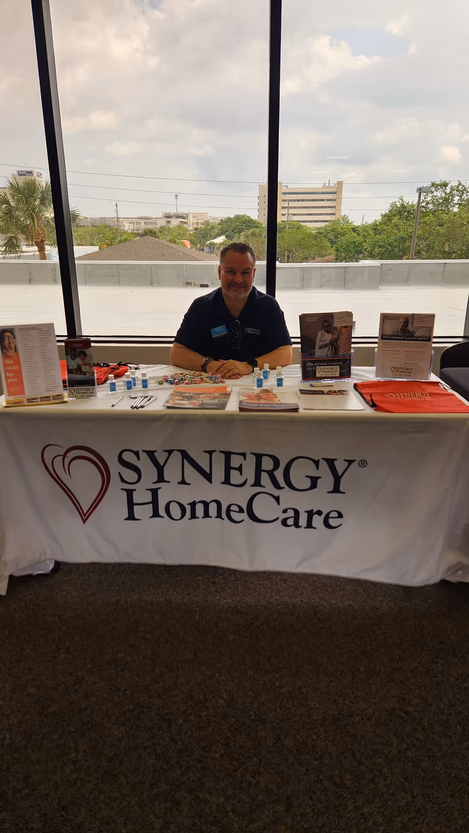 A man sits behind a SYNERGY HomeCare booth table with brochures indoors.