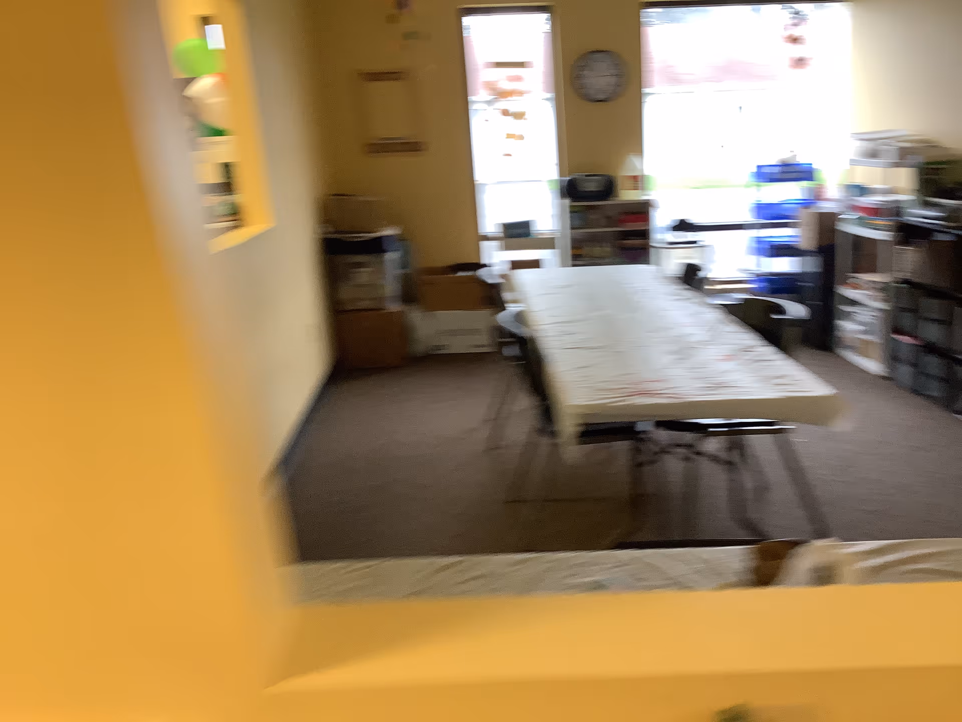 Blurred interior room with a long table, shelves, and storage clutter.