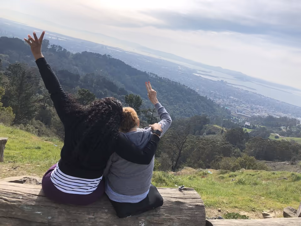 Two people sit on a log overlooking a hillside and distant cityscape.