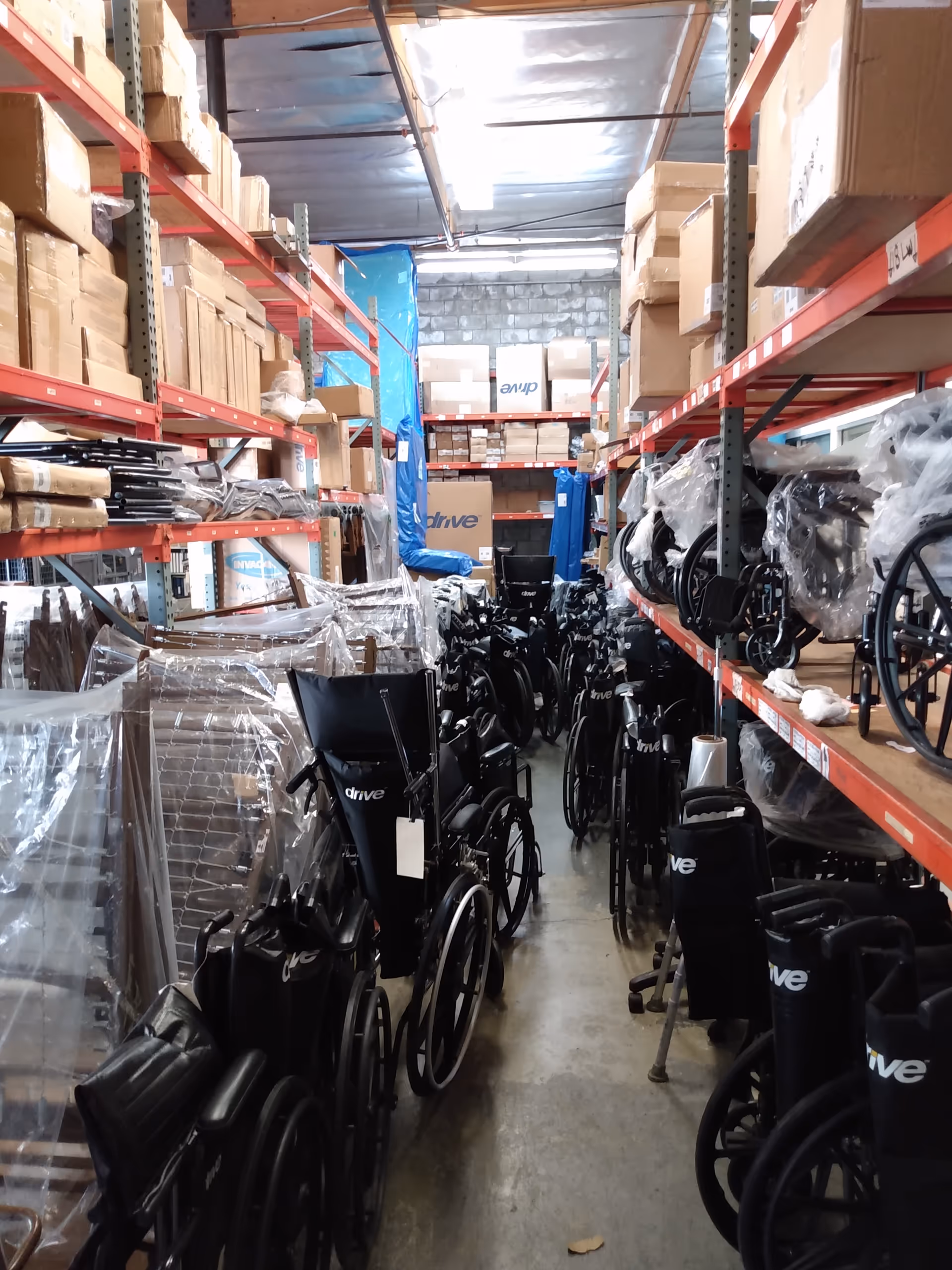 Narrow warehouse aisle filled with stacked wheelchairs and medical equipment on metal shelves.