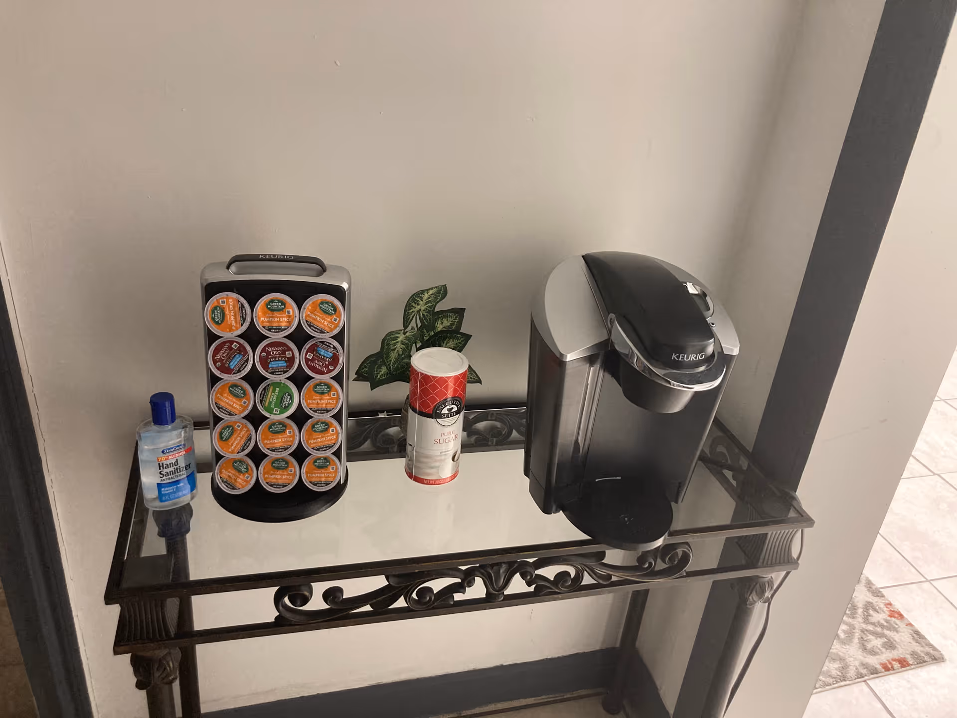 Coffee station with Keurig brewer, K-Cups rack, hand sanitizer, and a small plant on glass shelf.