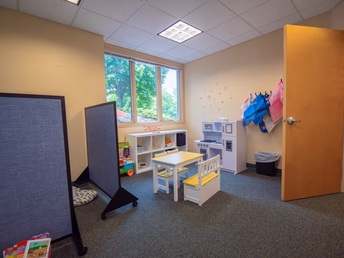 Bright interior playroom with a small table, chairs, toy kitchen, and cubbies by a window.