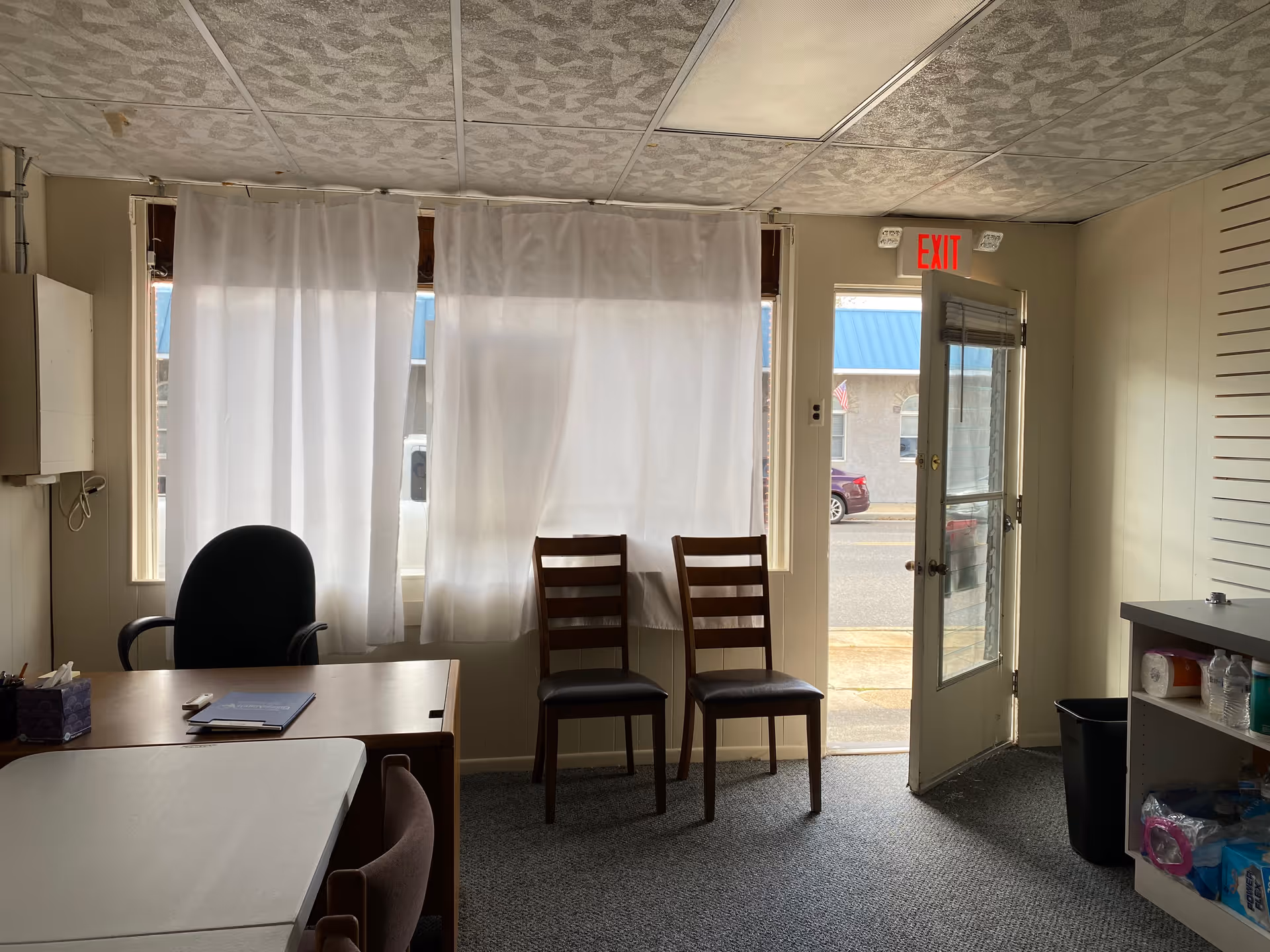 Office-like waiting area with desk, chairs, and curtained windows beside a door.