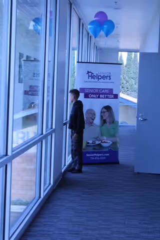 Indoor corridor with balloons and a Senior Helpers banner, two people standing by windows.