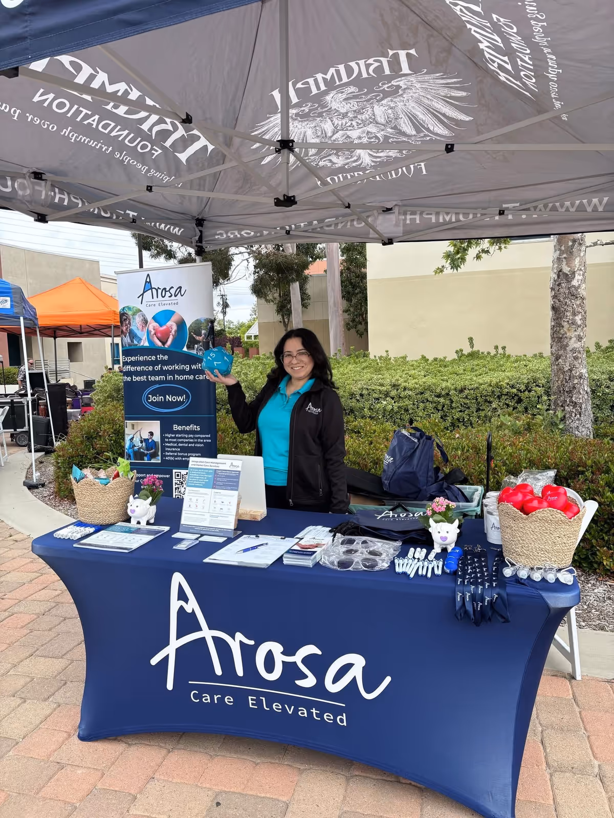 A staff member stands at an Arosa booth outdoors with banners and a table.