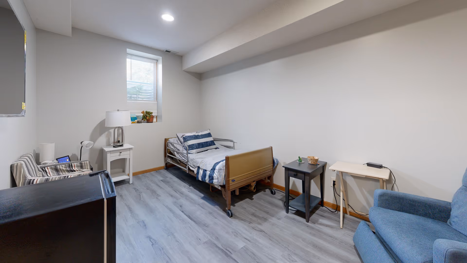 A small furnished senior living bedroom featuring a bed, sofa, nightstands, and a window.