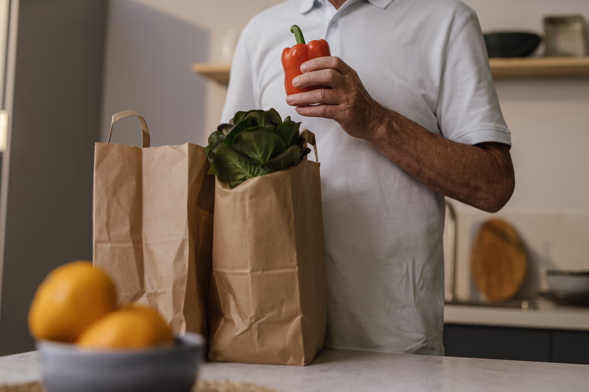 A person in a kitchen holds a red pepper beside grocery bags on the counter.