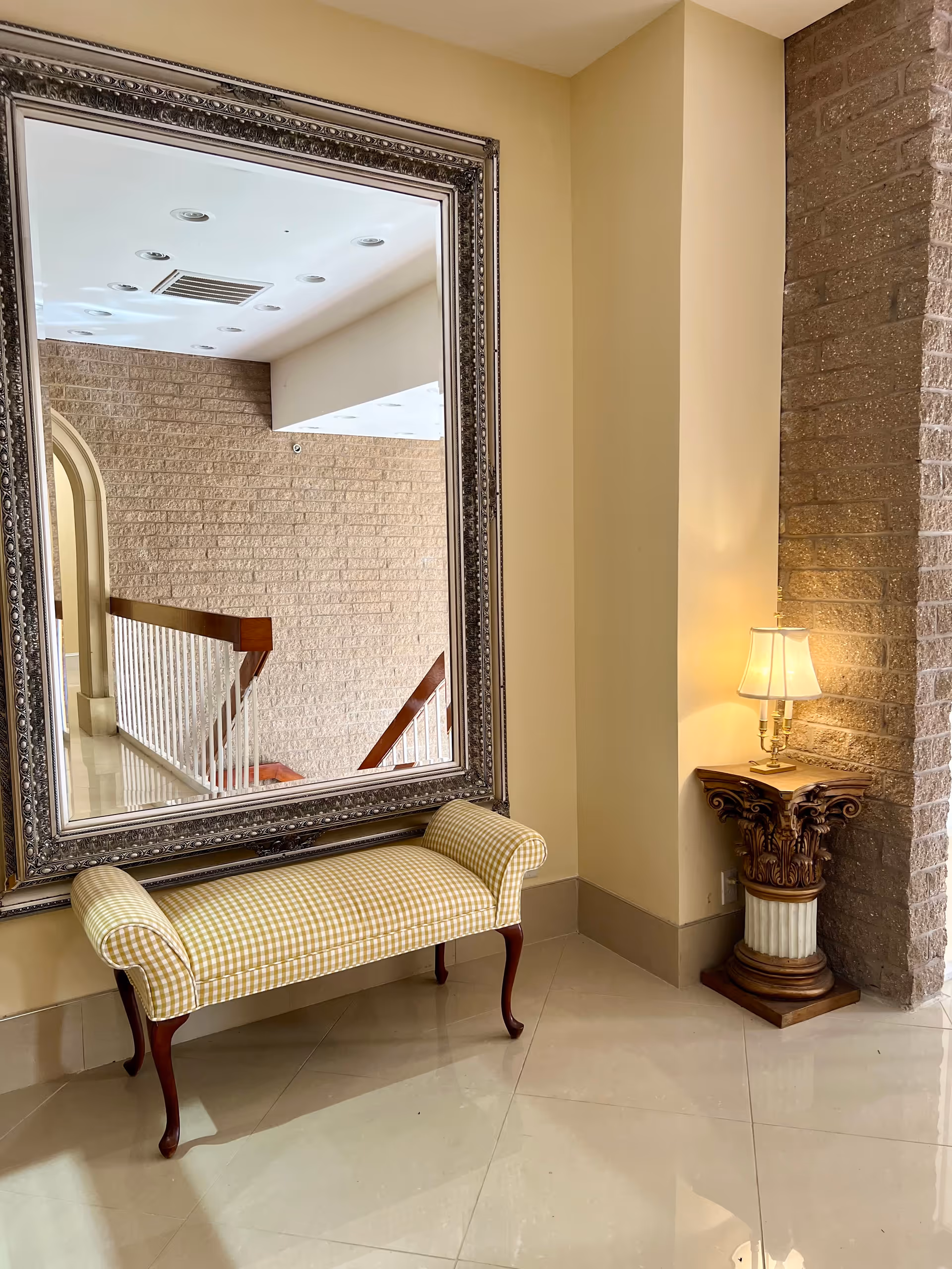 Ornate framed mirror over a checkered bench reflects a stairwell in a cream hallway, with lamp.