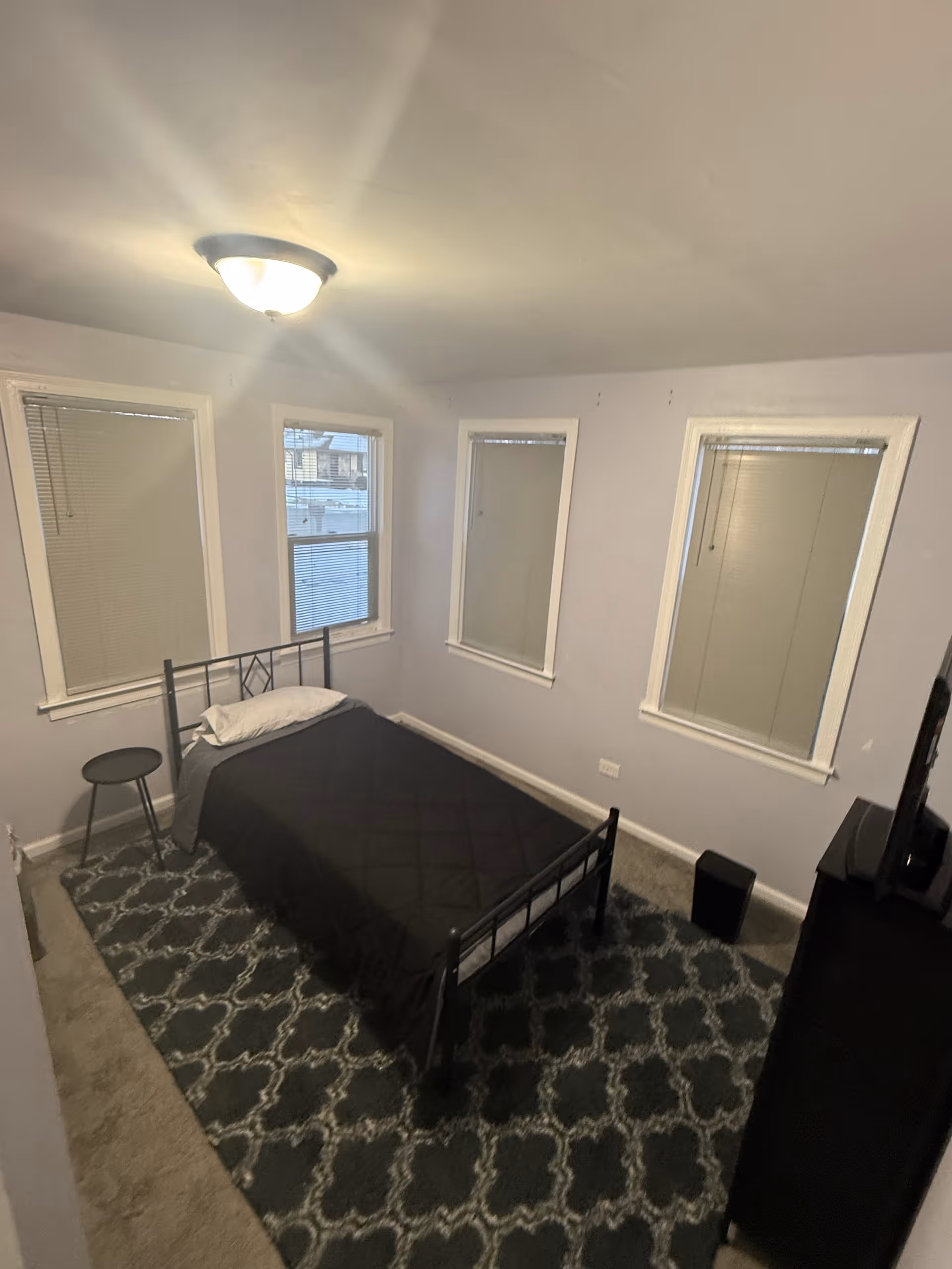 Small bedroom with metal bed, stool, rug, and four windows along gray walls.
