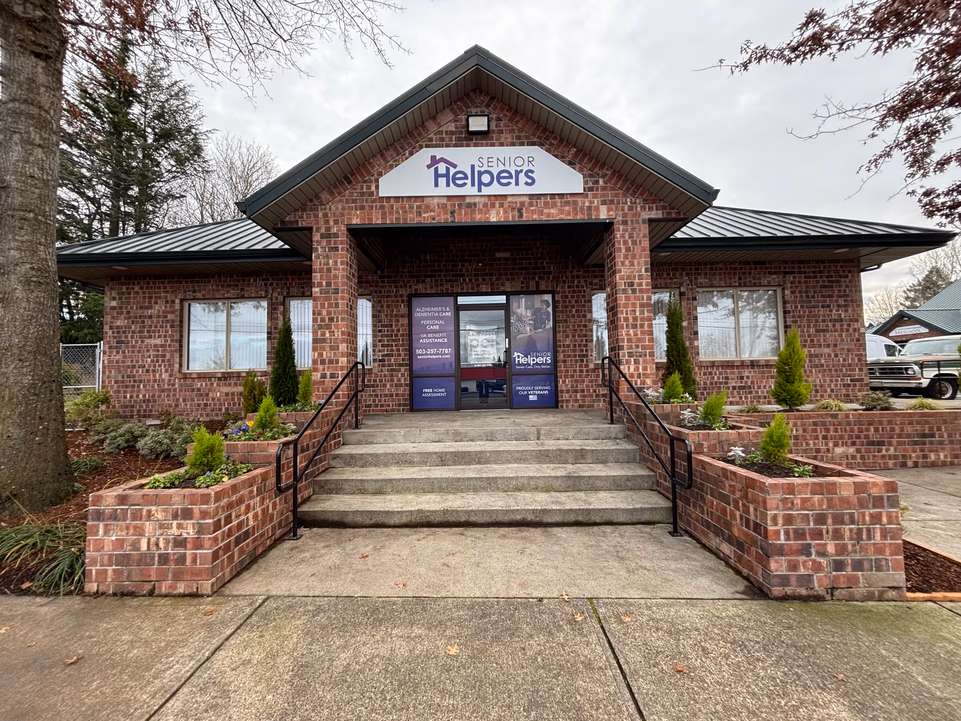 Front view of a brick Senior Helpers facility with stairs, railings, and landscaped planters.