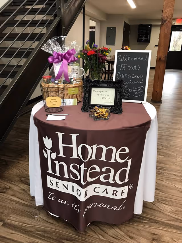 Indoor lobby table for Home Instead Senior Care with flowers, gifts, and chalkboard sign.