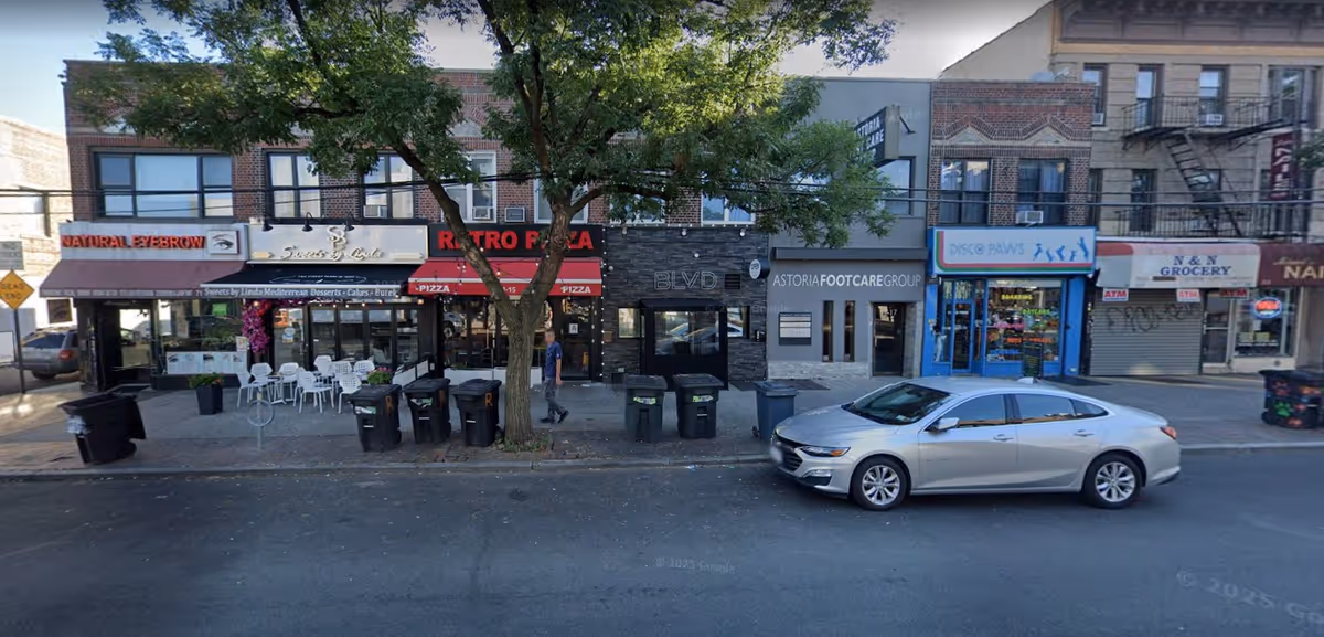 Street view with row of storefronts, outdoor seating, and a tree along the sidewalk.