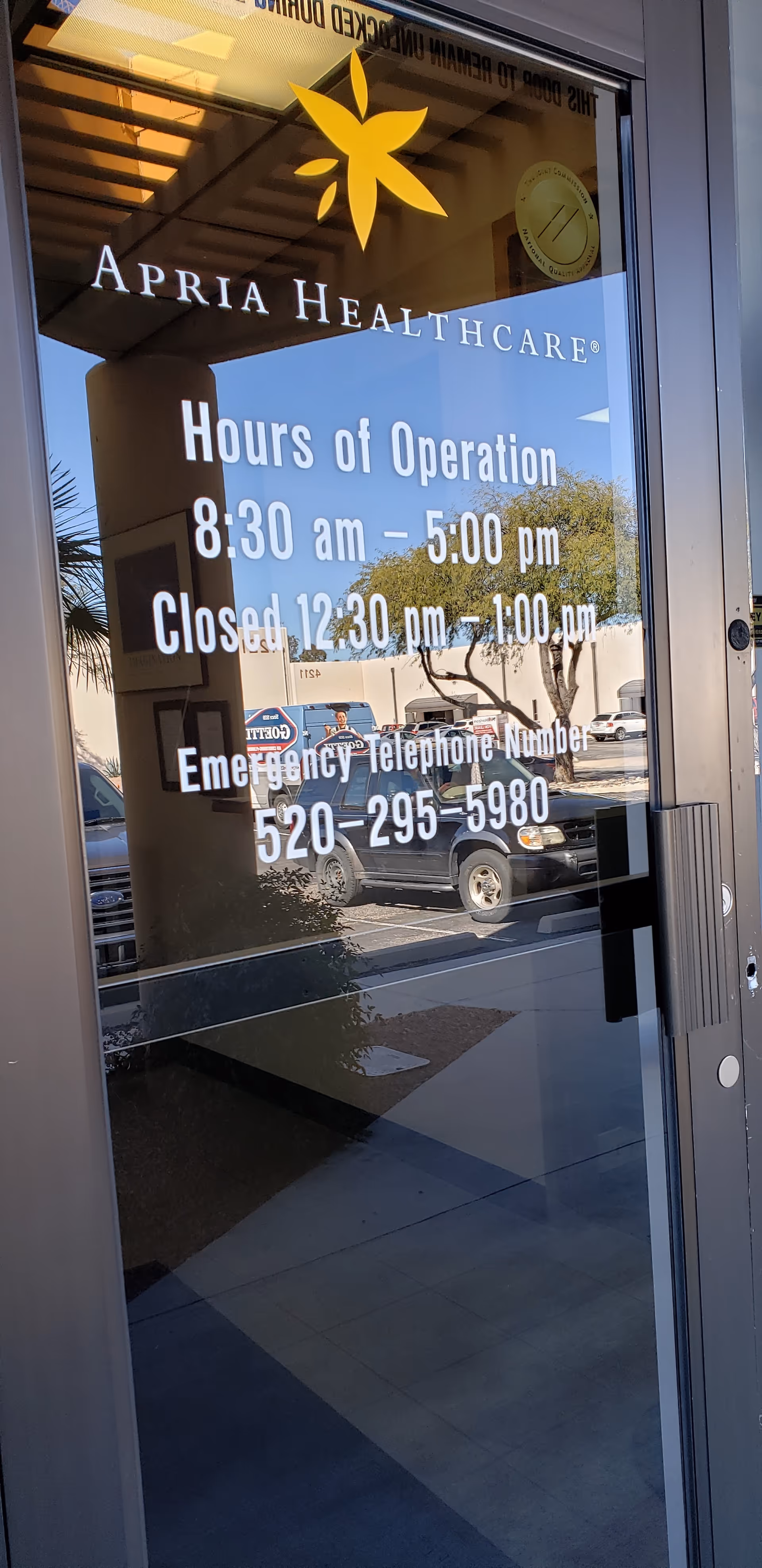 Glass storefront door shows Apria Healthcare hours and logo; reflection of a parked car outside.