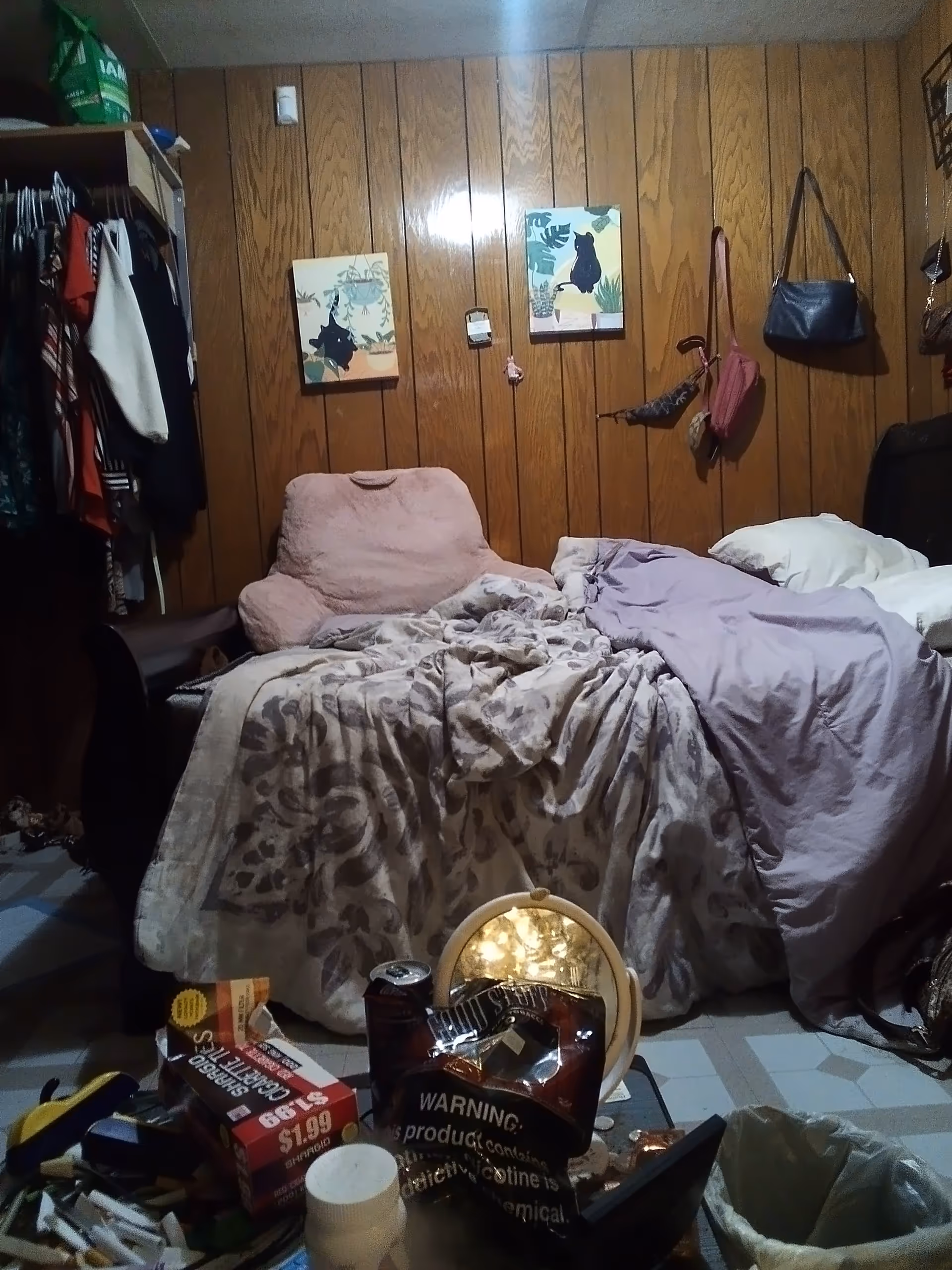 A cluttered senior living bedroom with a bed, clothing, and bags against wood panel walls.