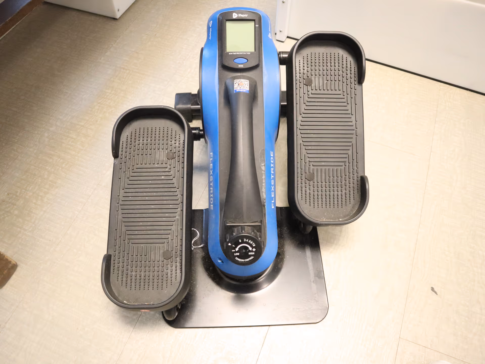 Blue exercise pedal machine with dual pedals sits on a beige tiled facility interior.