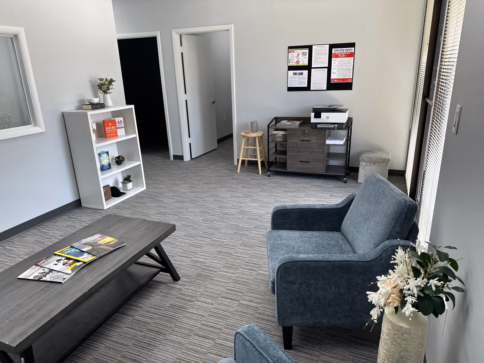 Neutral gray lounge in a senior facility with blue armchairs, coffee table, magazines, and a desk.
