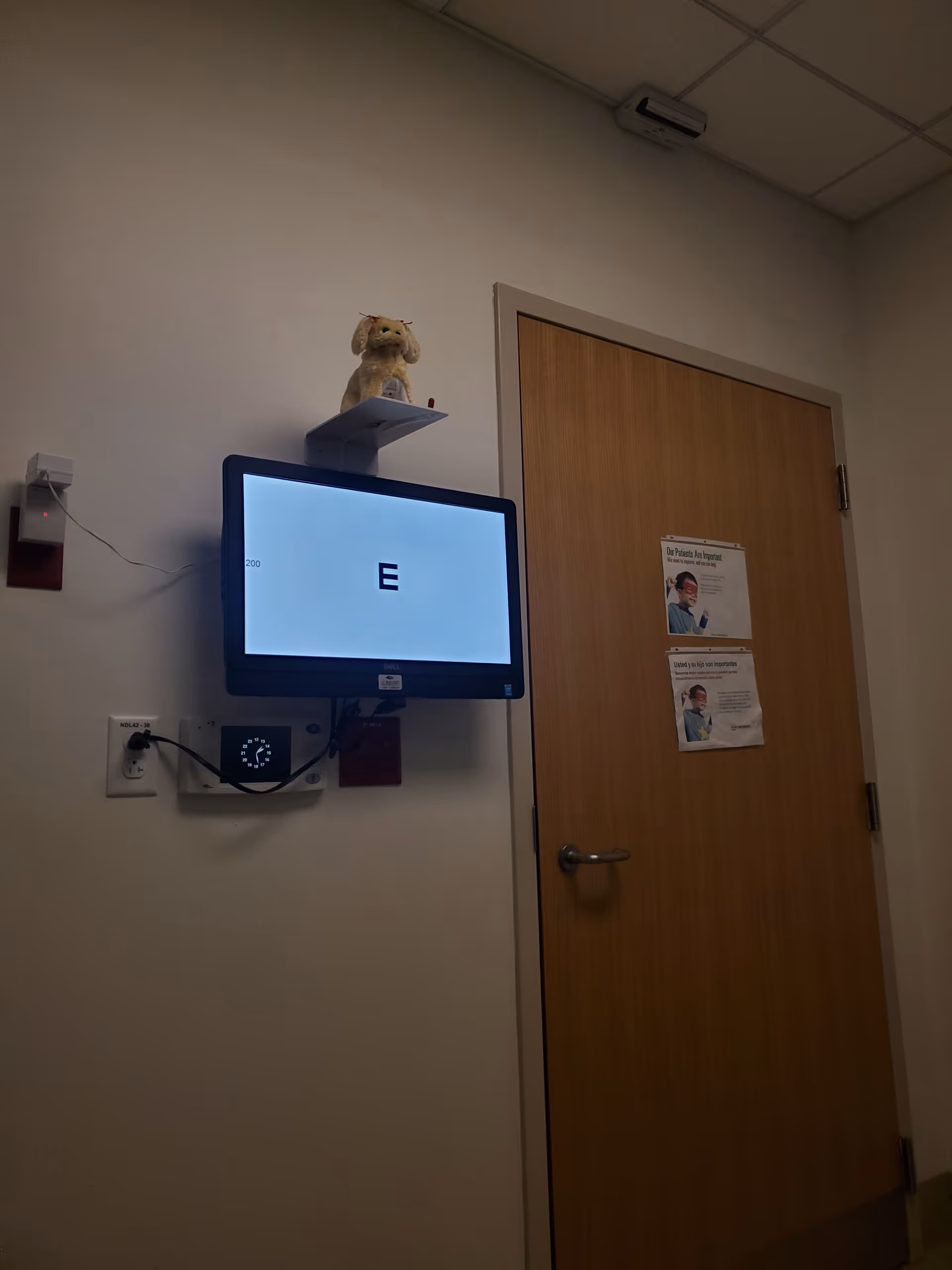 Interior hallway with wall-mounted monitor showing E, door, posters, and a plush toy on a shelf.
