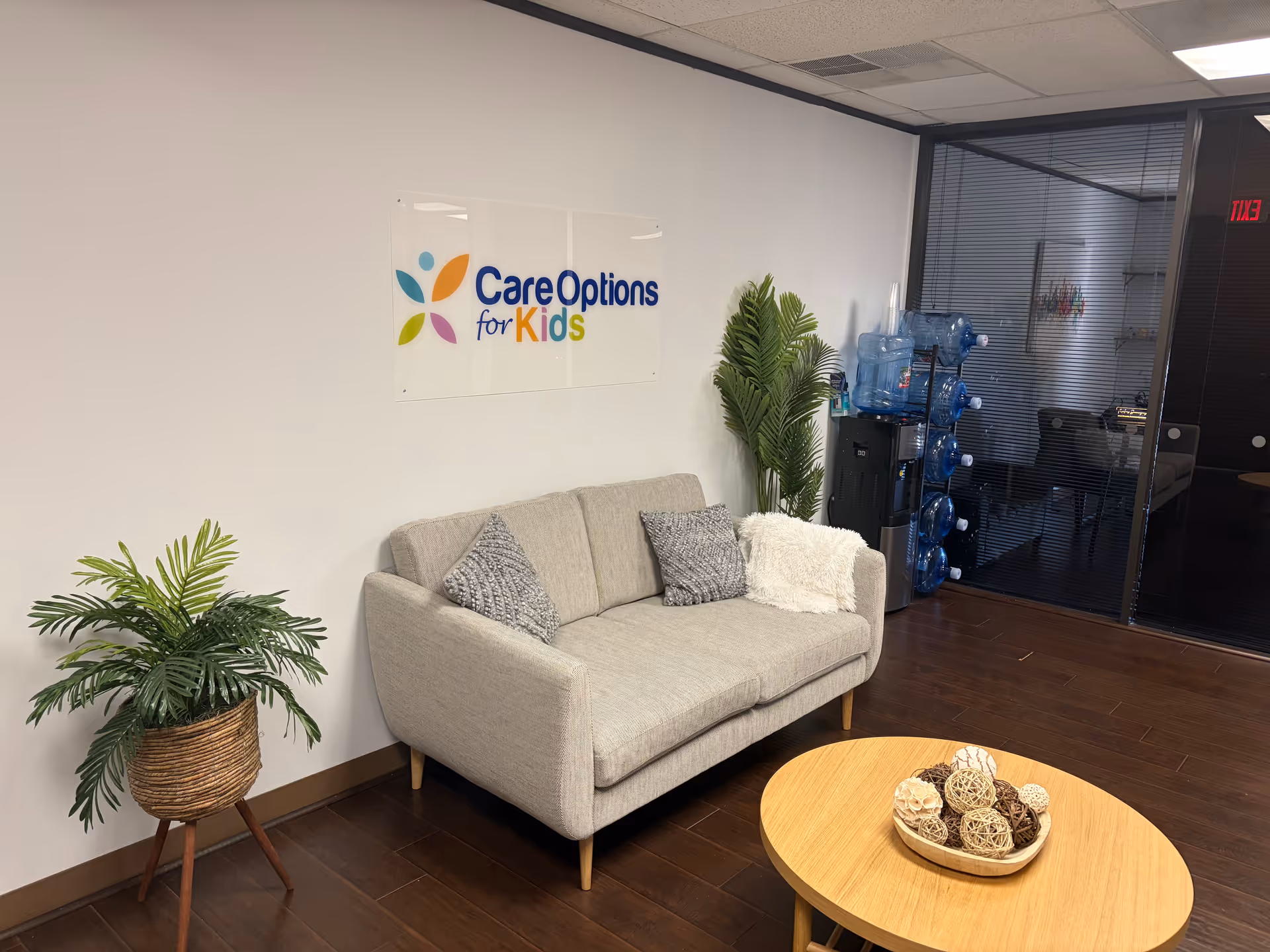 Grey sofa, plants, and a coffee table in a lobby with Care Options for Kids sign.