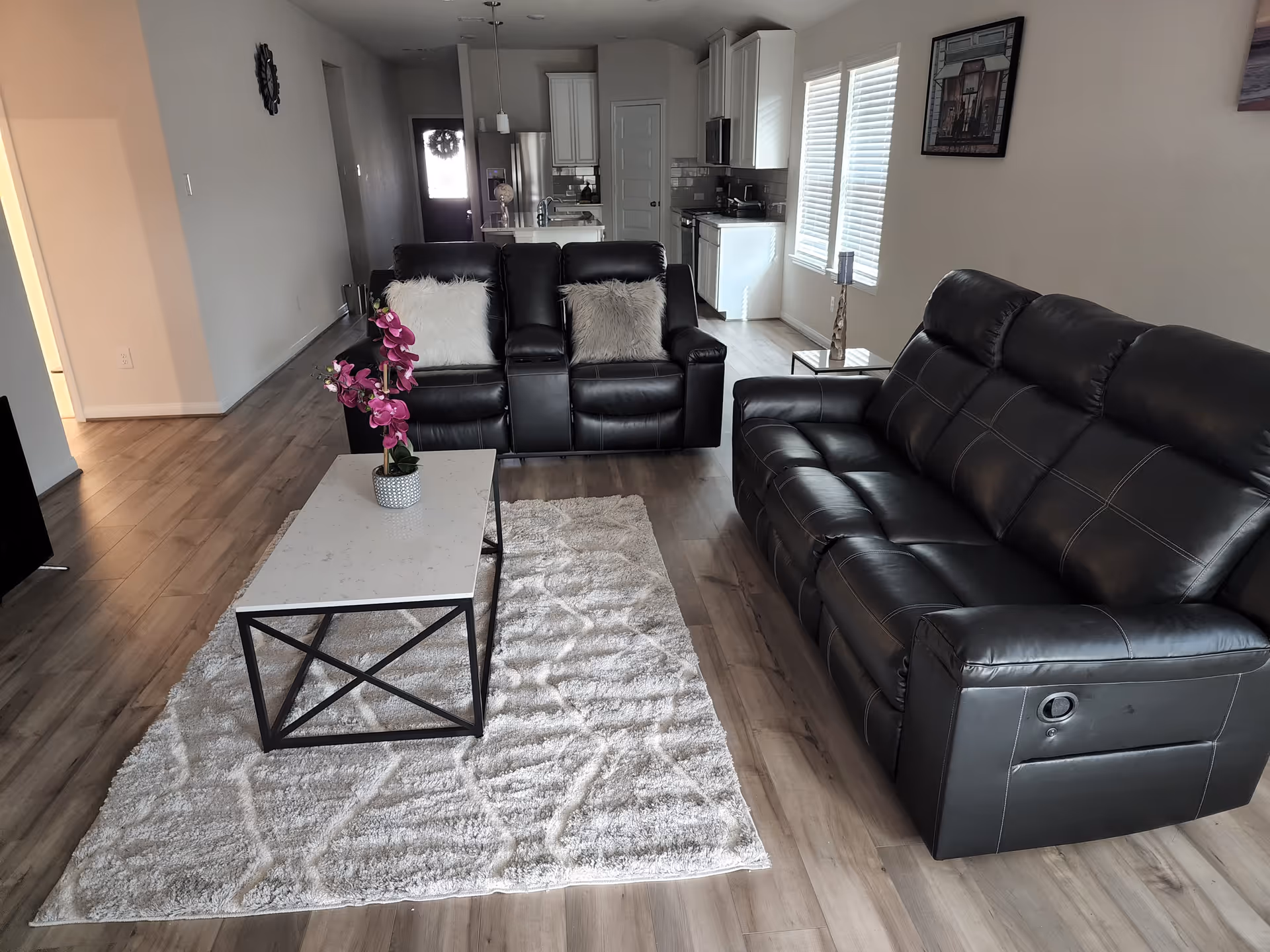 Open-concept living room with black leather sofas, coffee table, rug, and kitchen in background.