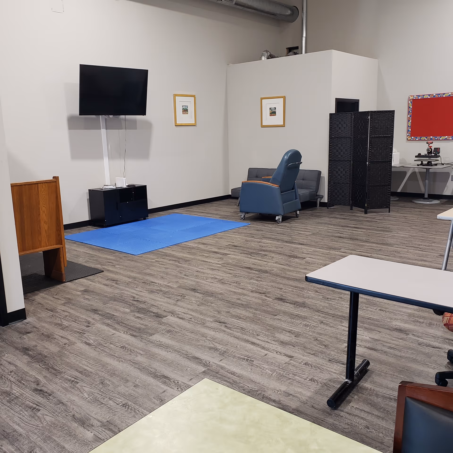 Senior living activity room with a blue recliner, TV, blue exercise mat and partition screens.