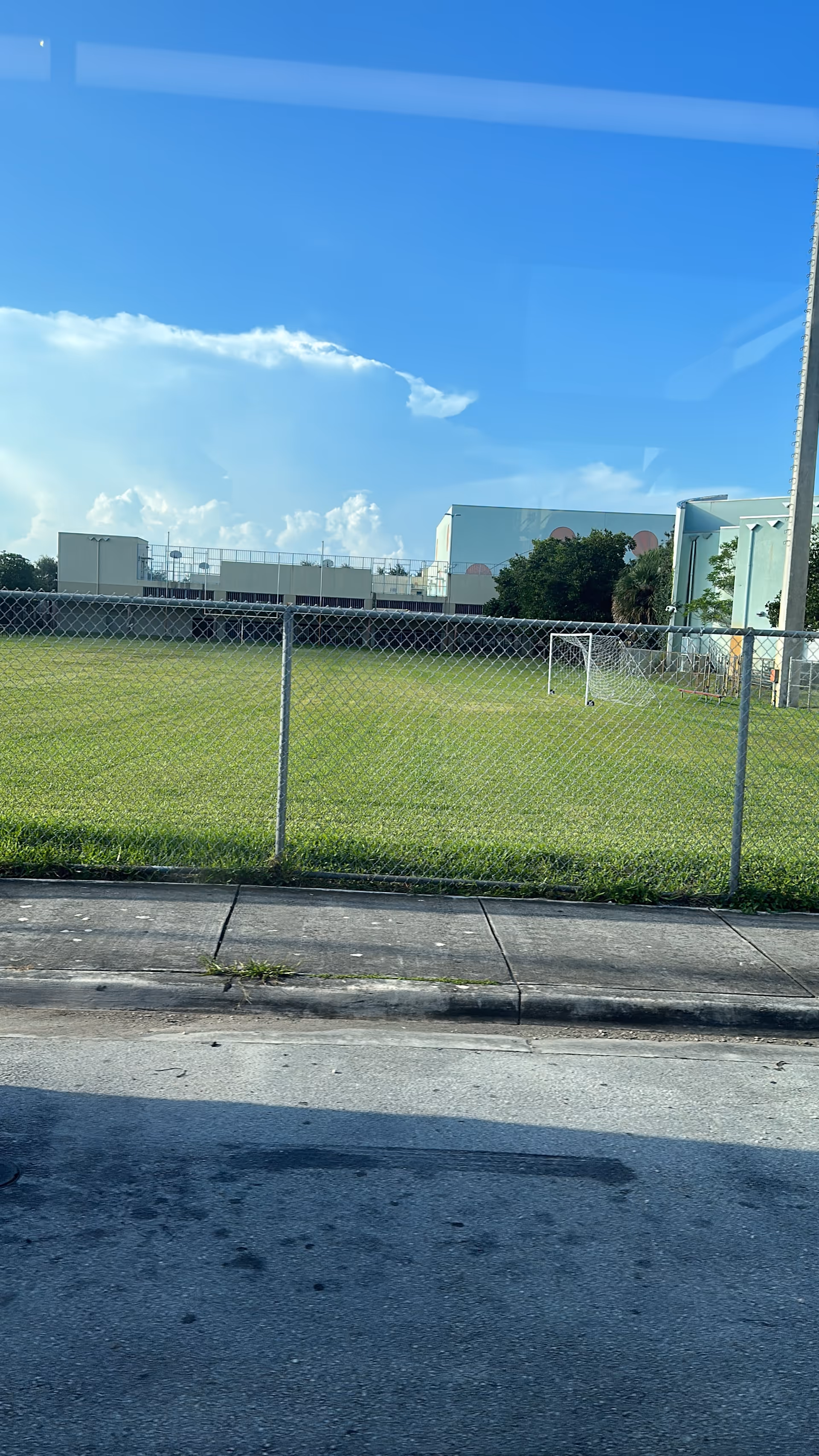 Outdoor grassy field with a small soccer goal, chain-link fence, and distant buildings.