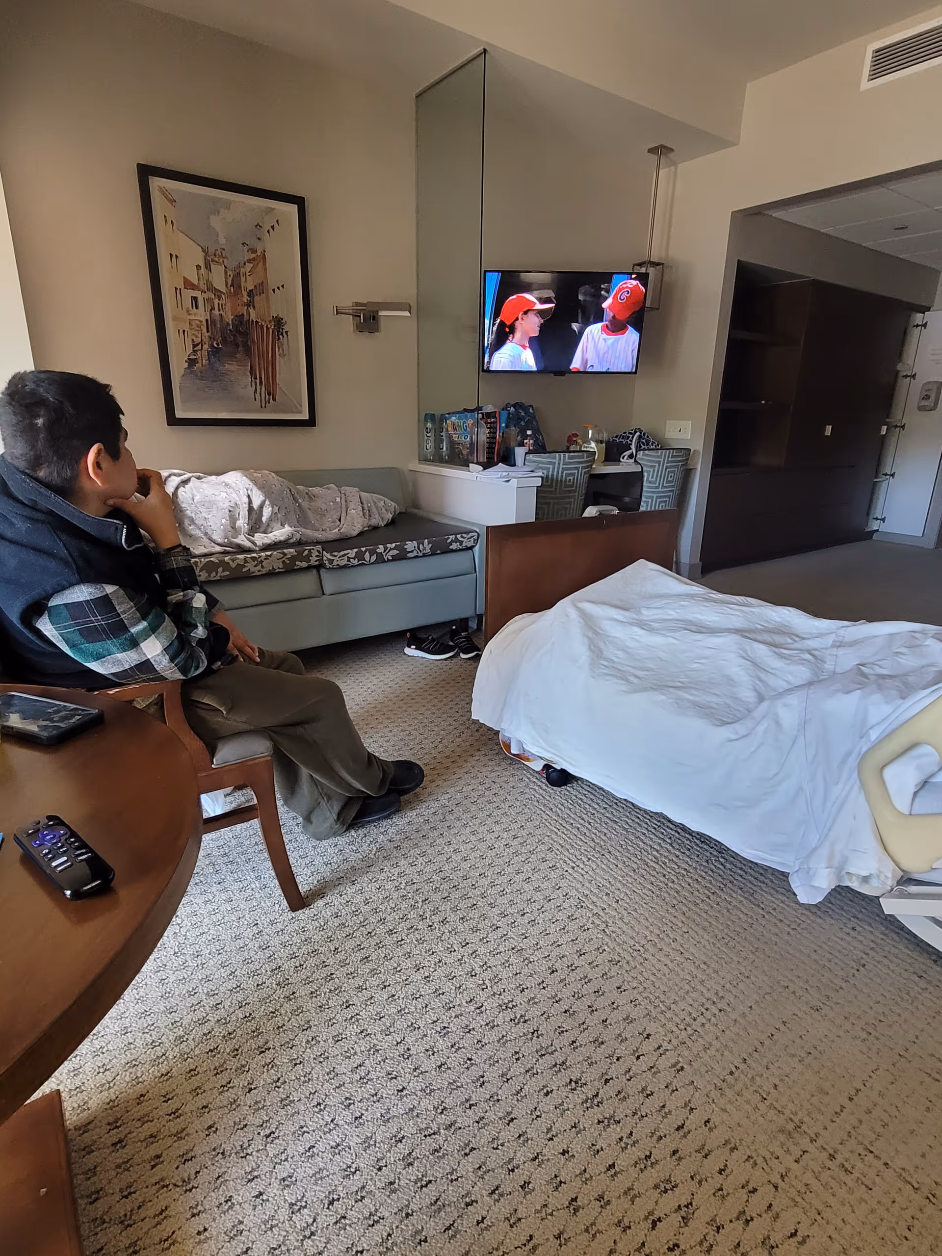 Hospice patient room with a bed, couch, TV, and a man seated nearby.