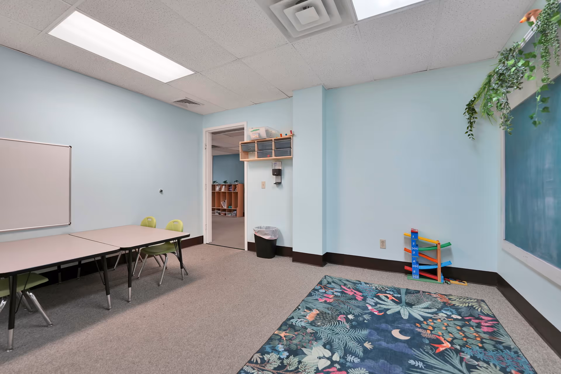 A light blue interior activity room with tables, chairs, a rug, and classroom supplies.