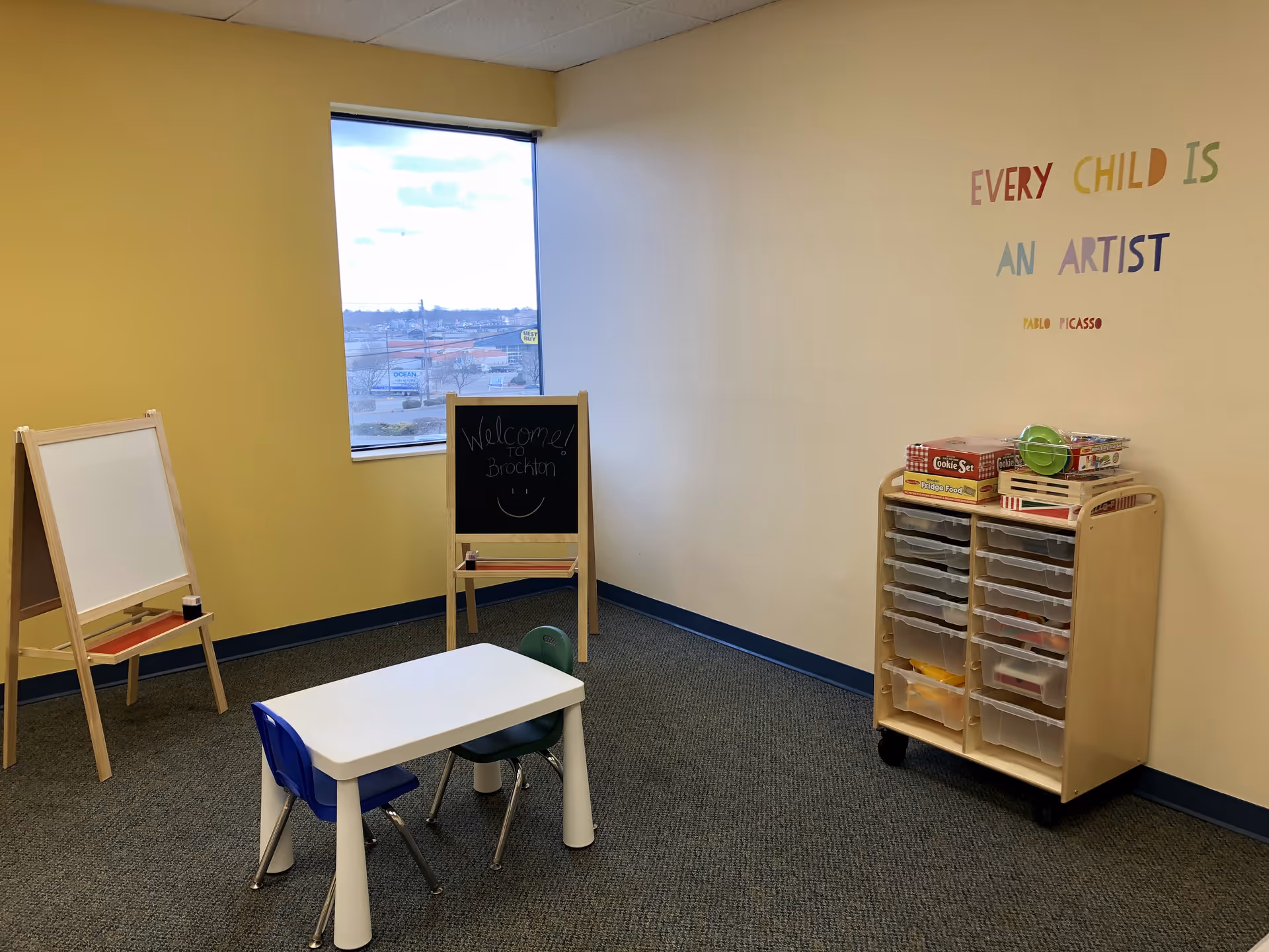 Small activity room with child-sized table, chairs, easels, chalkboard, and storage cart.