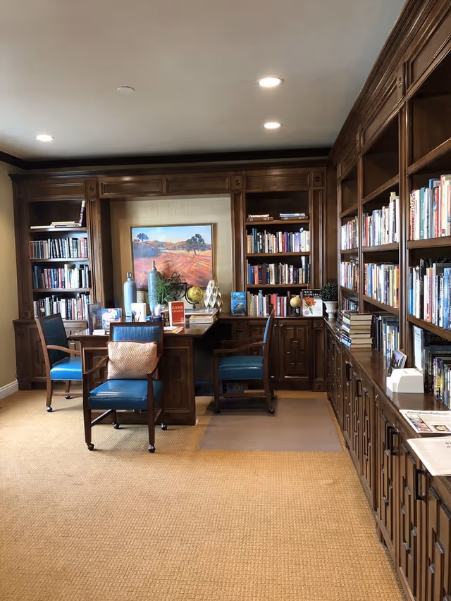 Cozy library study with dark wood built-in bookshelves, desks, blue chairs, and a landscape painting.