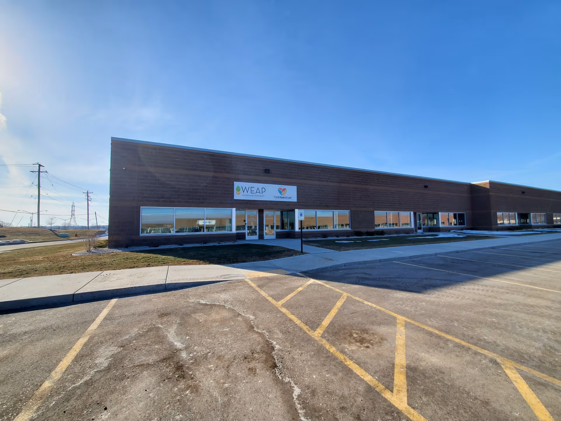 Brick one-story building with WEAP sign in Wisconsin, large windows facing sunny parking lot.